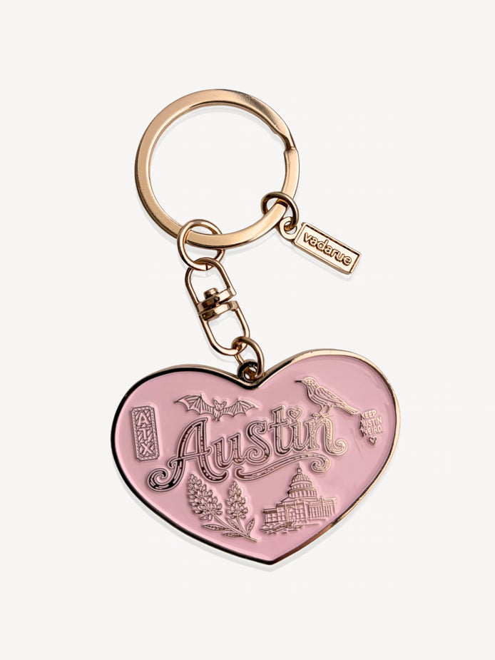 A pink heart-shaped keychain charm with 'Austin' and local landmarks engraved, attached to a rose gold split ring and keychain hardware on a light gray background.