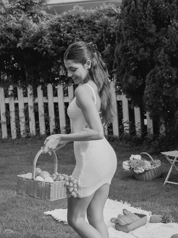 A woman in a white crochet mini dress stands in a grassy backyard holding a wicker basket of fruit and grapes, with a picnic blanket and baskets on the ground nearby. Photo is black and white.
