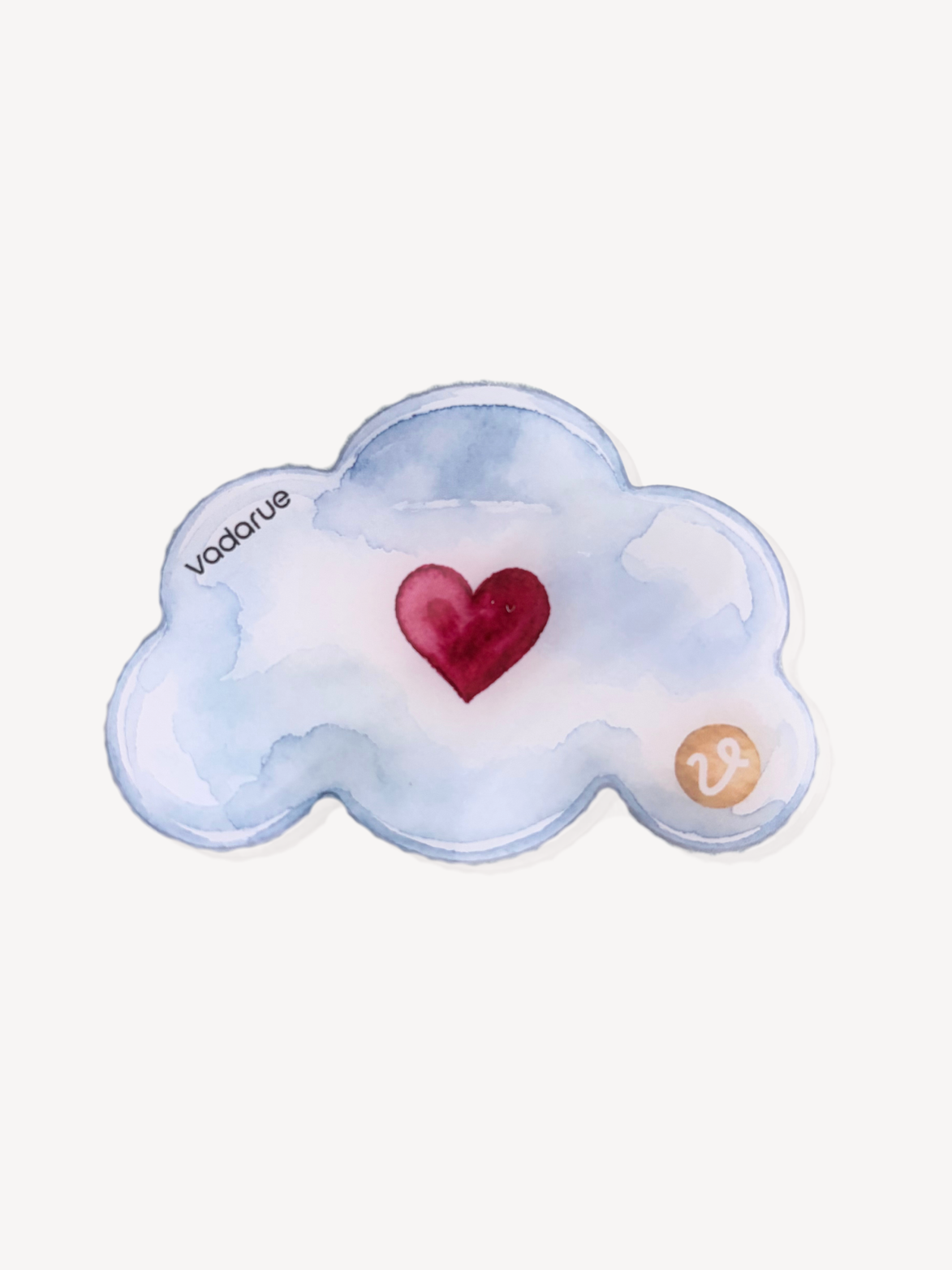 A watercolor-style cloud-shaped product in pale blue and white with a red heart in the center and a small gold monogram mark near the lower right, shown on a plain light background. The word 'vadorue' appears near the left edge of the cloud.