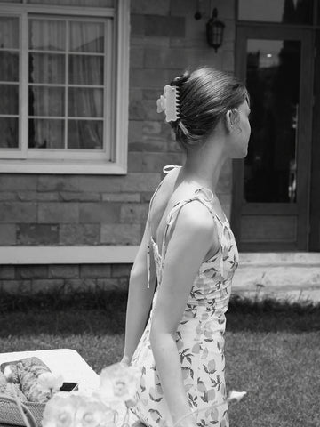 A woman in a sleeveless floral print dress stands outdoors in front of a house, shown from the back and side. The photo is in black and white, and a basket and table are partially visible in the foreground. 