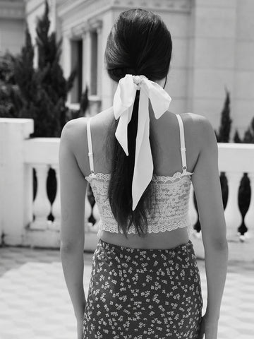 A woman viewed from behind wearing a white lace camisole top with a long white ribbon tied in her dark hair, paired with a dark floral skirt, set against an outdoor architectural background.