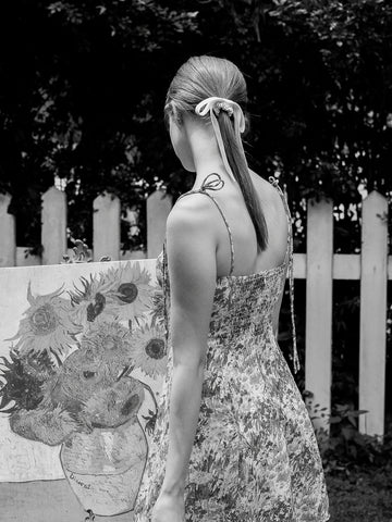 A woman seen from behind wearing a floral-patterned spaghetti-strap dress, standing outdoors next to a large sunflower painting in front of a white picket fence. photo is black and white.