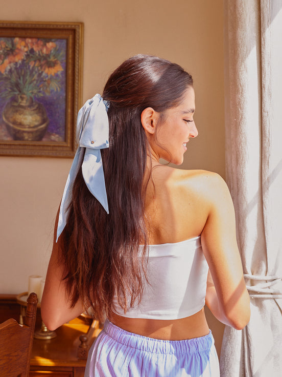 A woman with long dark brown hair wearing a white strapless tube top and light purple shorts, seen from behind as she looks over her shoulder. Her hair is styled with a large light blue silk ribbon bow.