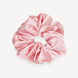 A light pink silk hair scrunchie with a small gold logo plate closure, shown on a clean off-white background.