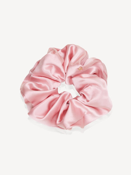 A light pink silk hair scrunchie with a small gold logo plate closure, shown on a clean off-white background.