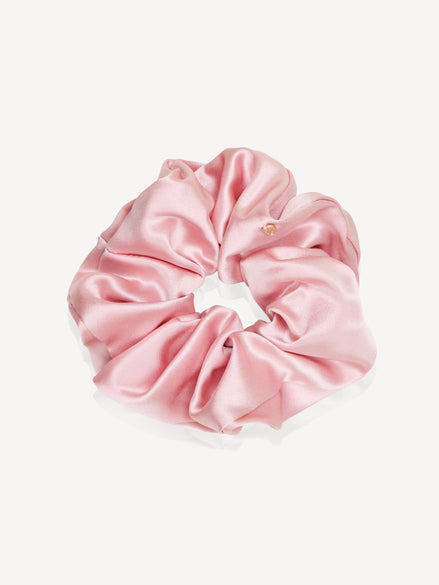 A light pink silk hair scrunchie with a small gold logo plate closure, shown on a clean off-white background.
