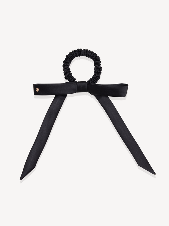 A black silk scrunchie hair tie with a wide silk bow and pointed ribbon ends, photographed flat against a light gray background.