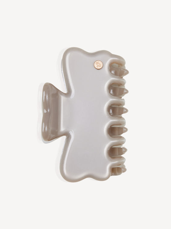 A glossy, light silver cellulose acetate hair claw clip with a small gold circular logo accent on the upper right, shown on a clean off-white background.