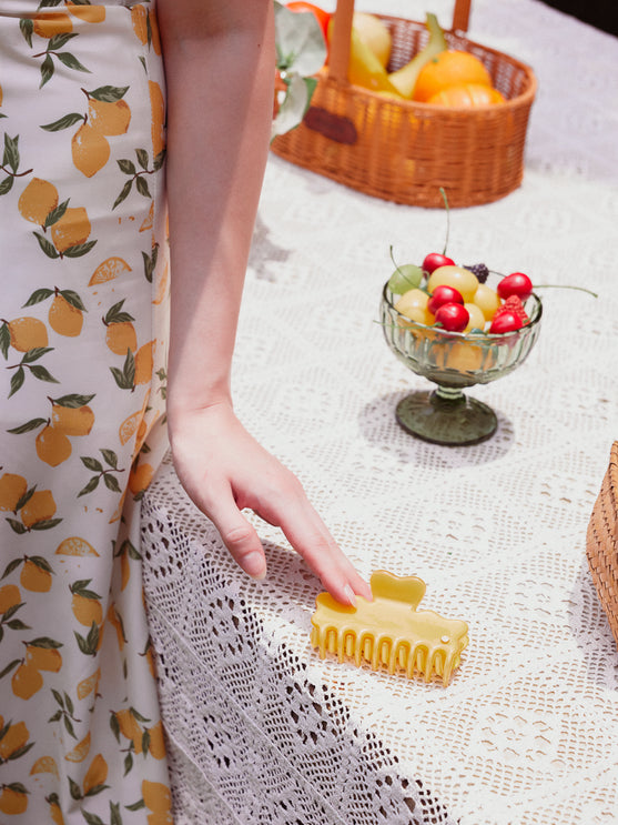 A person wearing a cream dress with a yellow lemon print is shown from the side, touching a small yellow cellulose acetate hair claw clip on a white lace tablecloth. A glass bowl of fruit and a wicker basket are in the background.