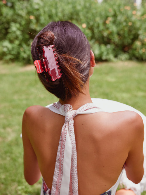 A woman seen from behind outdoors, wearing a white halter dress with a tie-back and a large red cellulose acetate claw hair clip holding her hair in a low bun.