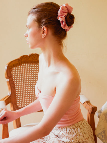 A woman in profile wearing a light pink strapless top and a textured light pink skirt, with her hair styled in a bun and adorned with a large pink silk scrunchie. She is seated in a wooden cane-back chair against a warm beige wall.
