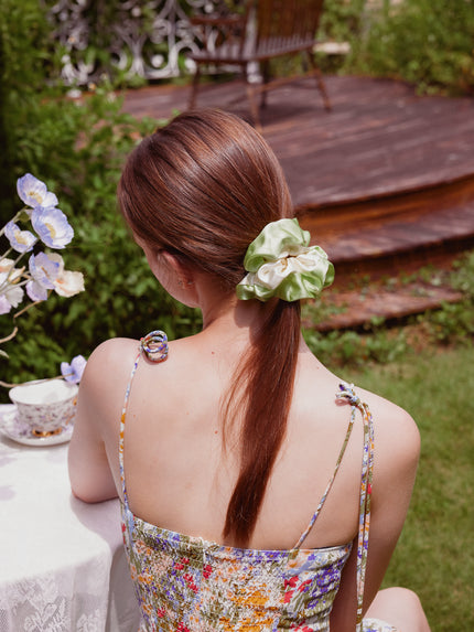A woman viewed from behind in an outdoor garden setting, wearing a light green silk scrunchie with a cream bow to tie her brown hair in a ponytail. She is wearing a colorful floral spaghetti-strap dress.