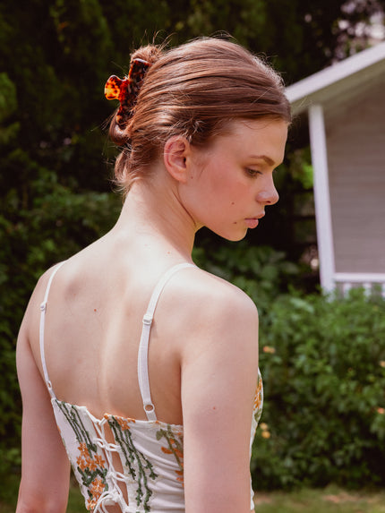 A woman outdoors wearing a white spaghetti-strap dress with a floral print and lace-up back, styled with a tortoise cellulose acetate hair clip.