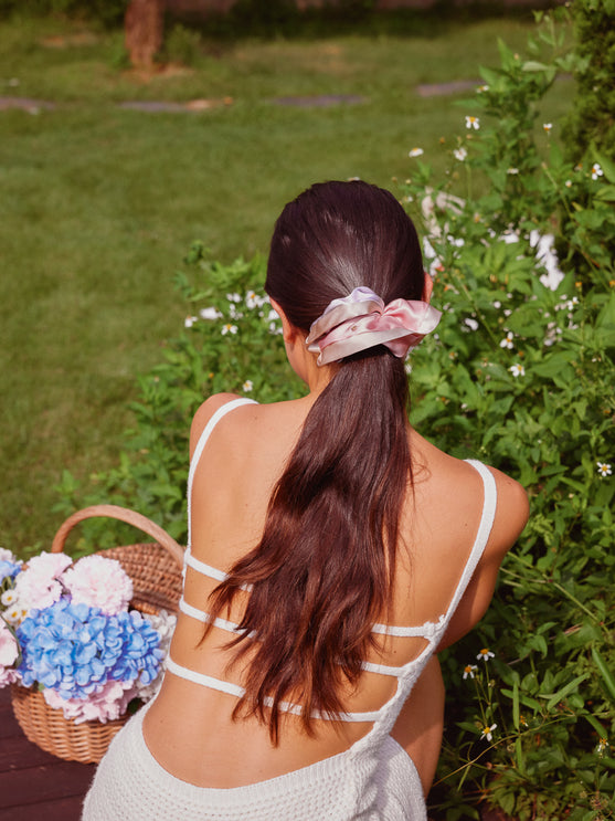 A woman viewed from behind wearing a white strappy camisole and a pink silk scrunchie, sitting outdoors in a garden next to a basket of blue and pink hydrangeas.