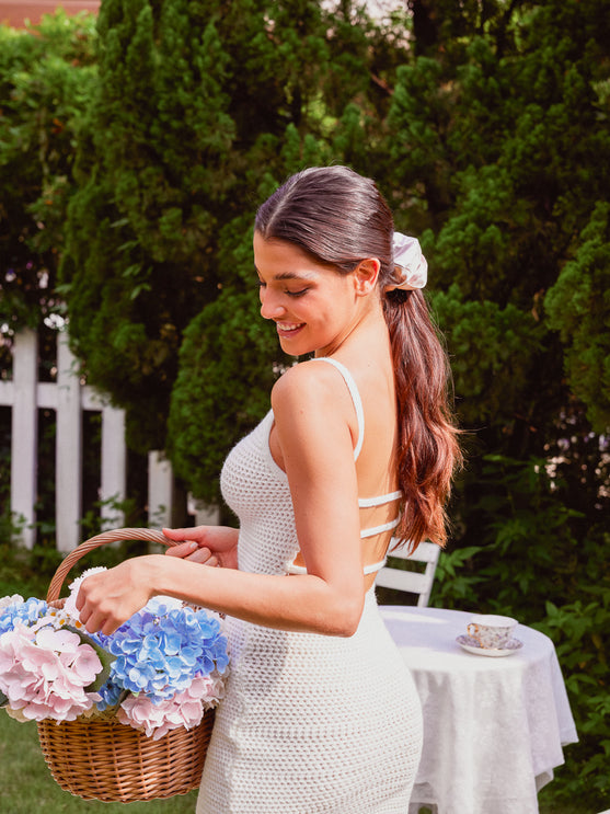 A woman in a white crochet dress holding a wicker basket of pink and blue hydrangeas, standing outdoors in front of green trees and a white table.