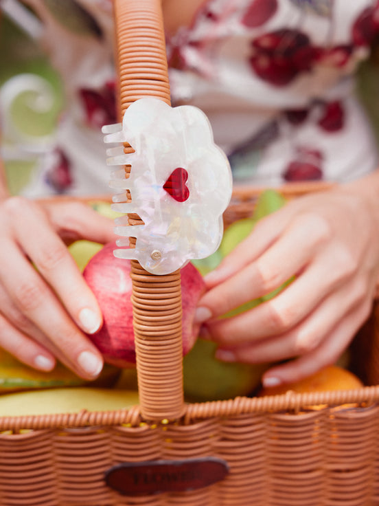 A close-up lifestyle shot of a person's hands arranging fruit inside a wicker basket, with a translucent white cloud-shaped cellulose acetate hair claw clip featuring a small red heart accent hanging from the basket's handle.